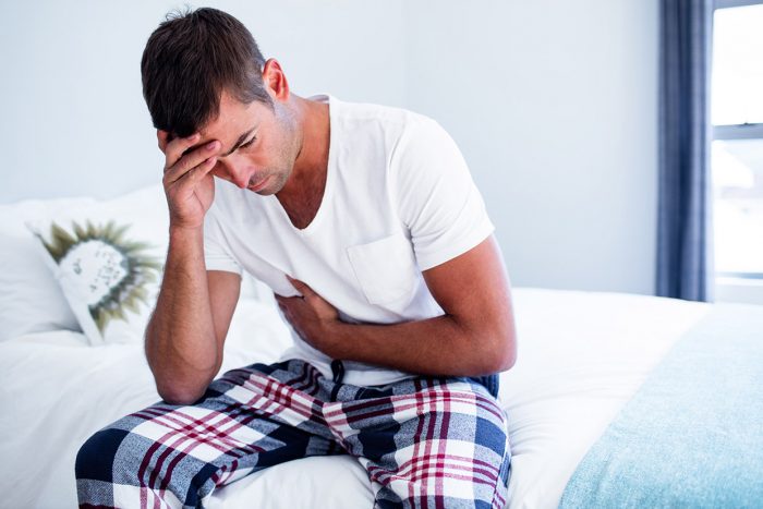 Young man sitting with stomach pain on bed in bedroom